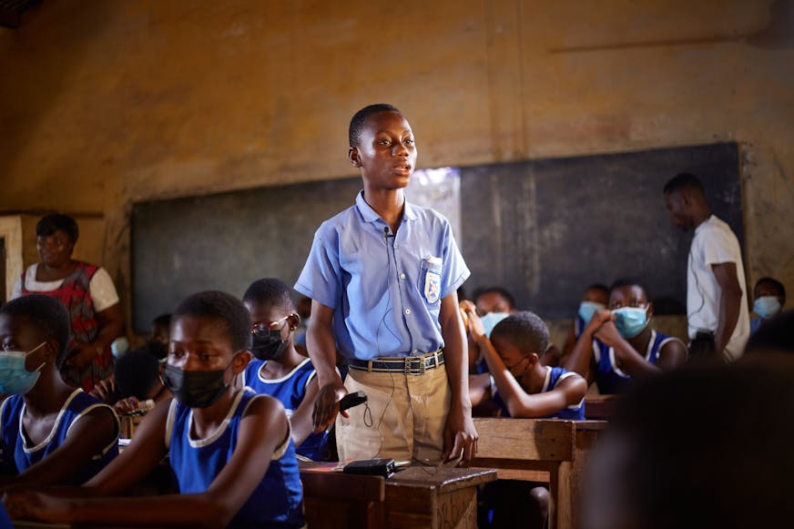 Kinderrechtenclubs - een jongen in een blauw uniform staat voor zijn bureau op een school in Ghana.